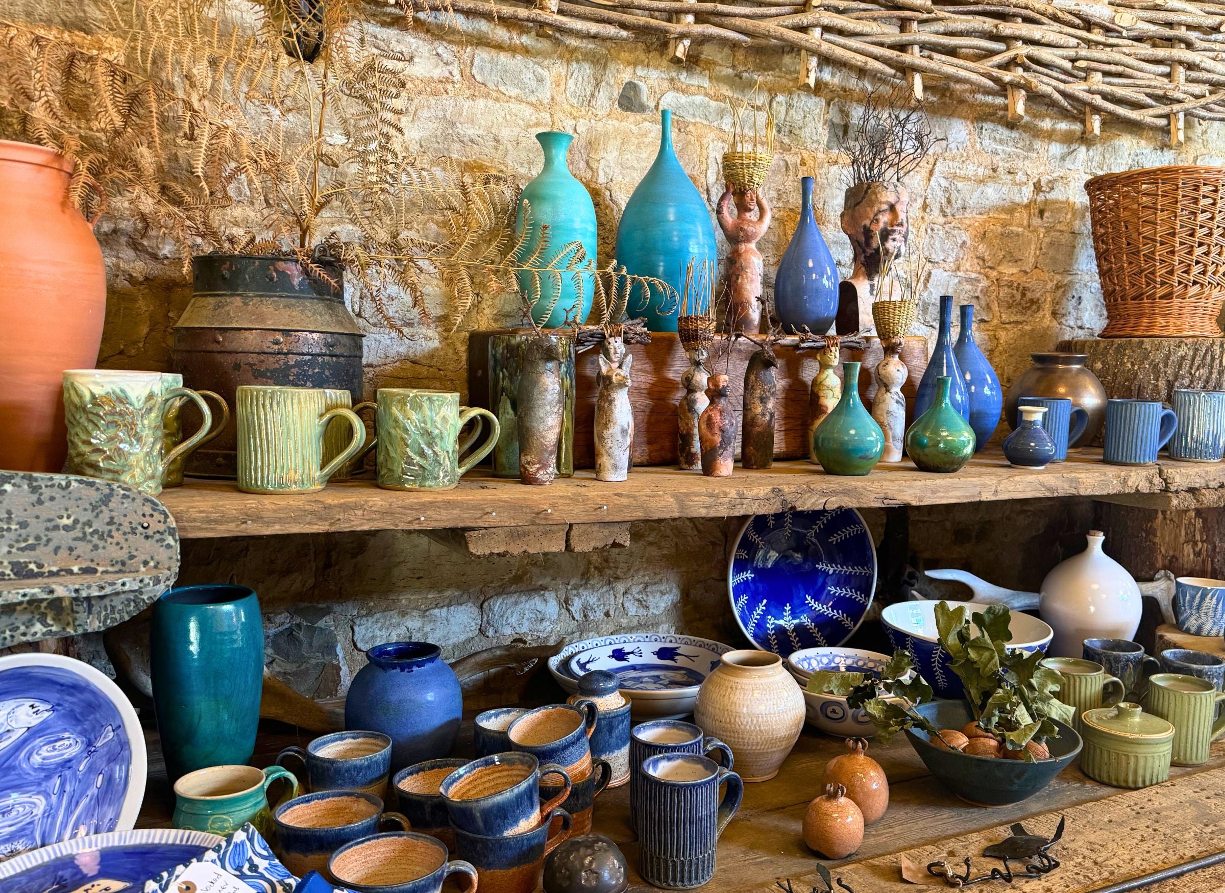 Collection of ceramic and pottery items on wooden shelves against a stone wall.